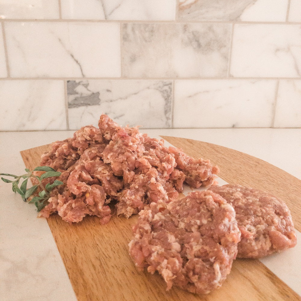 Beautifully displayed breakfast sausage not yet in casings on a cutting board with herbs and white marble tile background.
