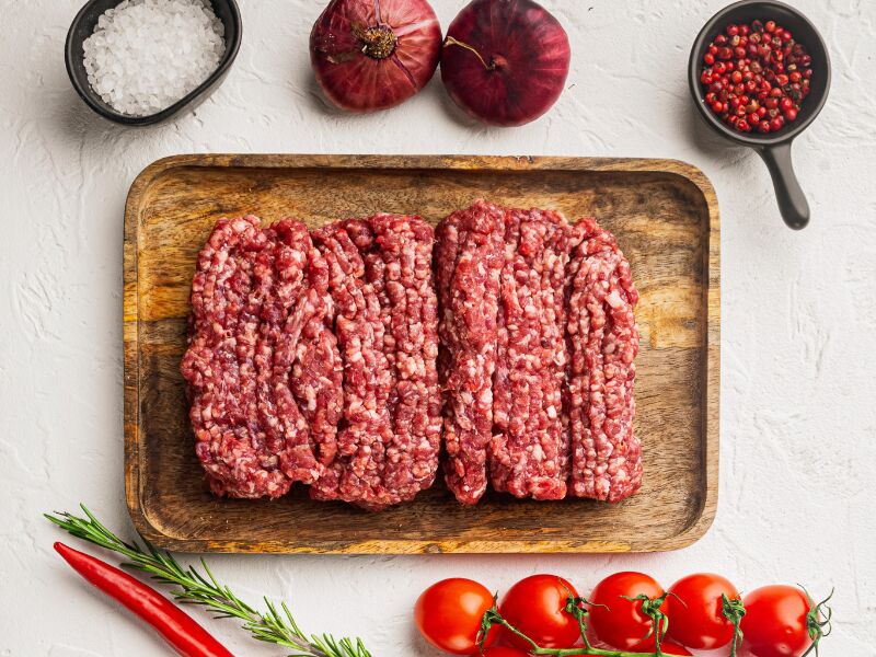 Ground lamb meat laid on a cutting board with whole on the vine tomatoes, whole red cayenne pepper, sprig of rosemary, two red onions, and bowls of coarse salt and black pepper.